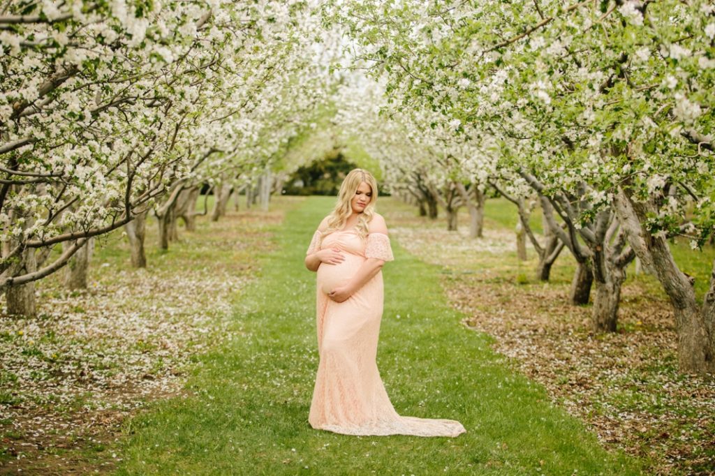 woman wearing pink dress with curled blonde hair looking over shoulder spring apple blossoms maternity session