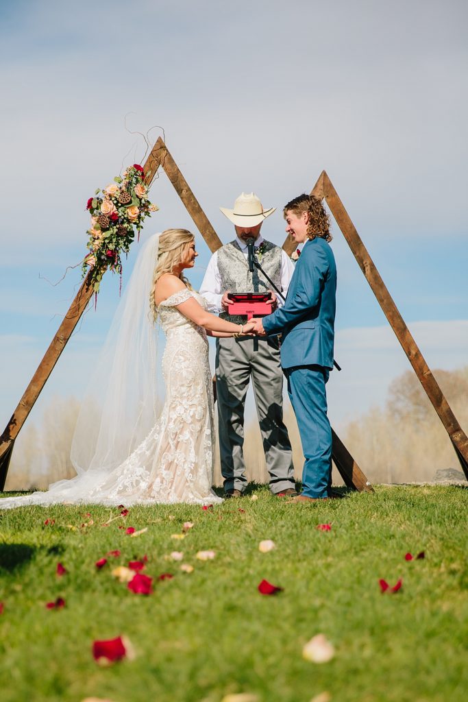 bride and groom standing under their triangle wedding arch in Pocatello Idaho as their officiant marries them during their summer wedding