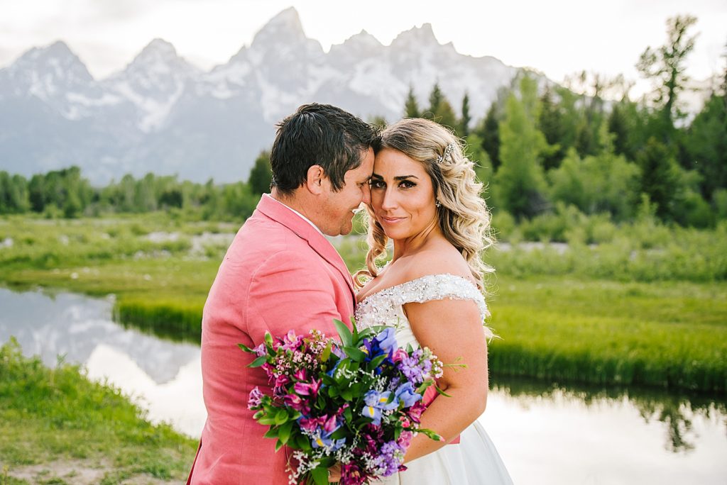 Grand Teton bride and groom standning in front of a river as the groom embraces the woman as he wears a pink suit for his wedding