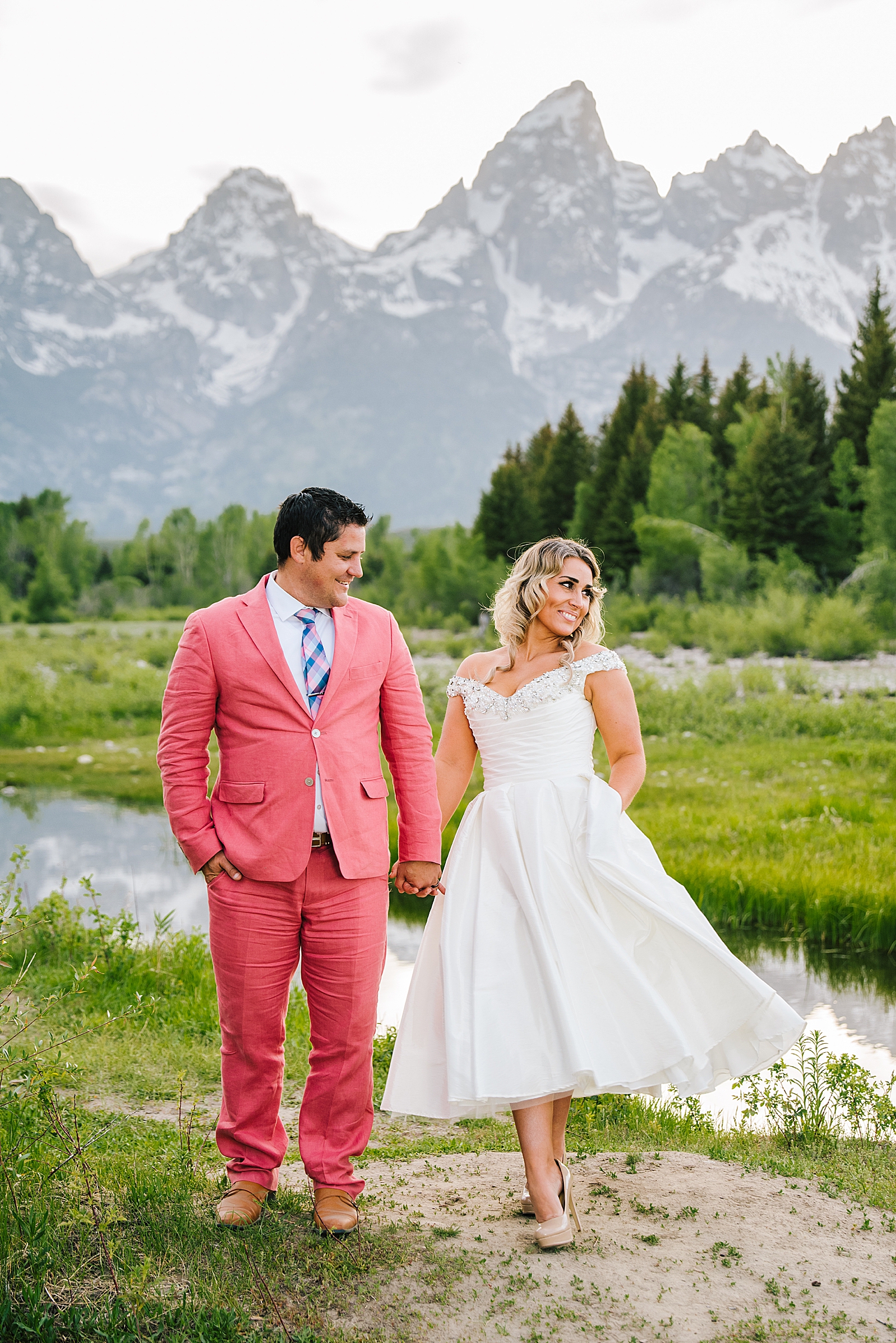 groom in a pink suit holds hands with his bride and walks along the river bed of the Teton's Snake River captured by Jackson Hole wedding photographer