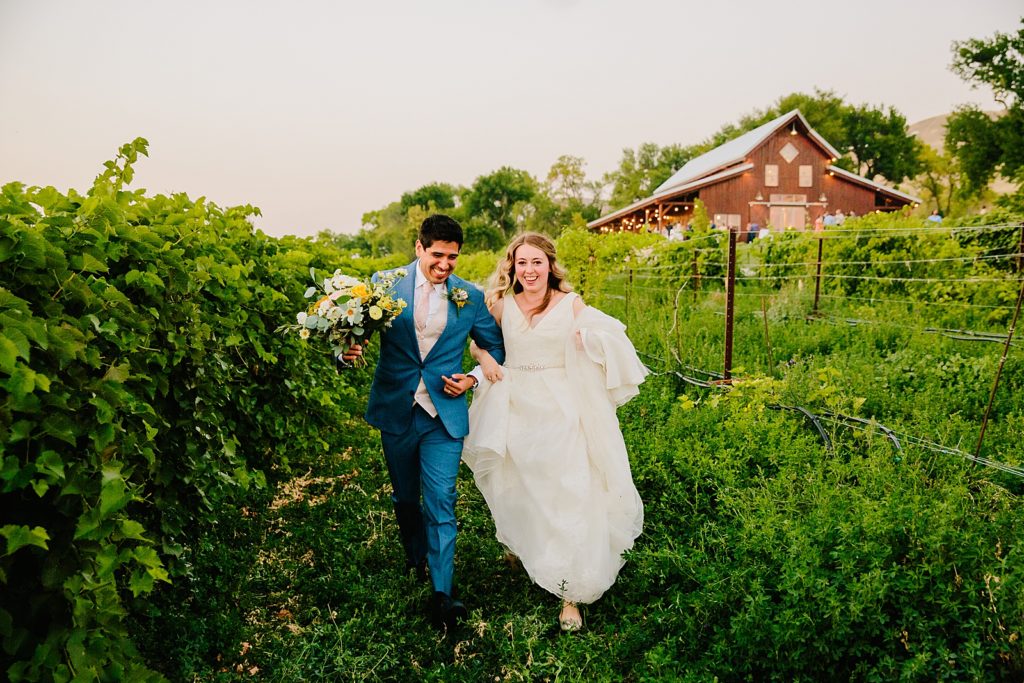bride and groom walking together outside of a barn ina green field with vineyards in Pocatello Idaho