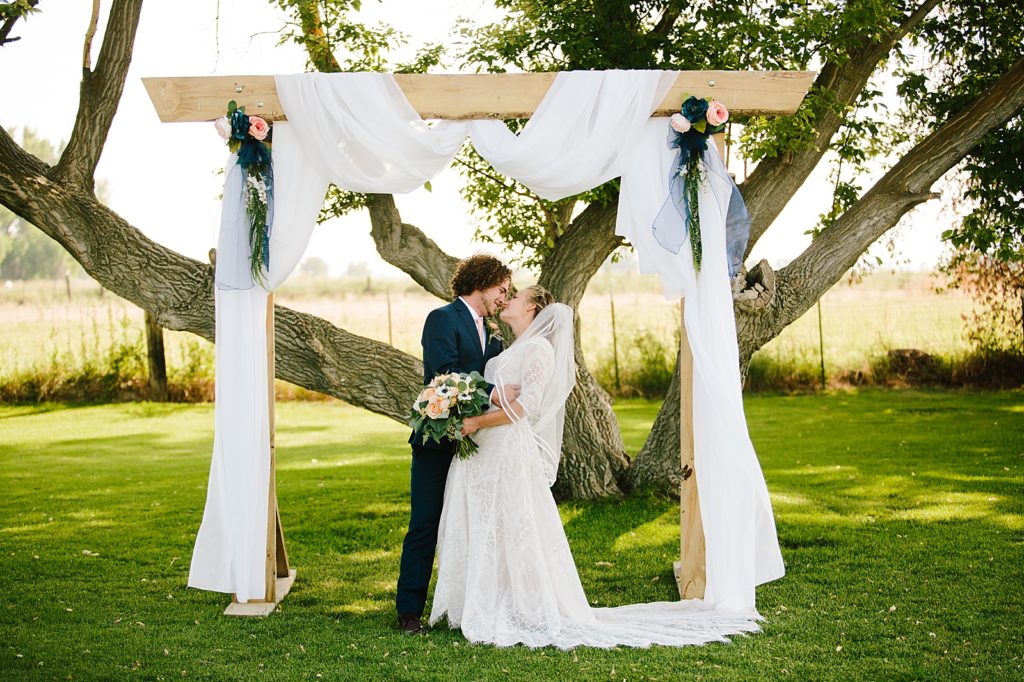 bride and groom standing together under their wedding arch together as they rest their foreheads together as the sun shines through the trees on their Idaho wedding