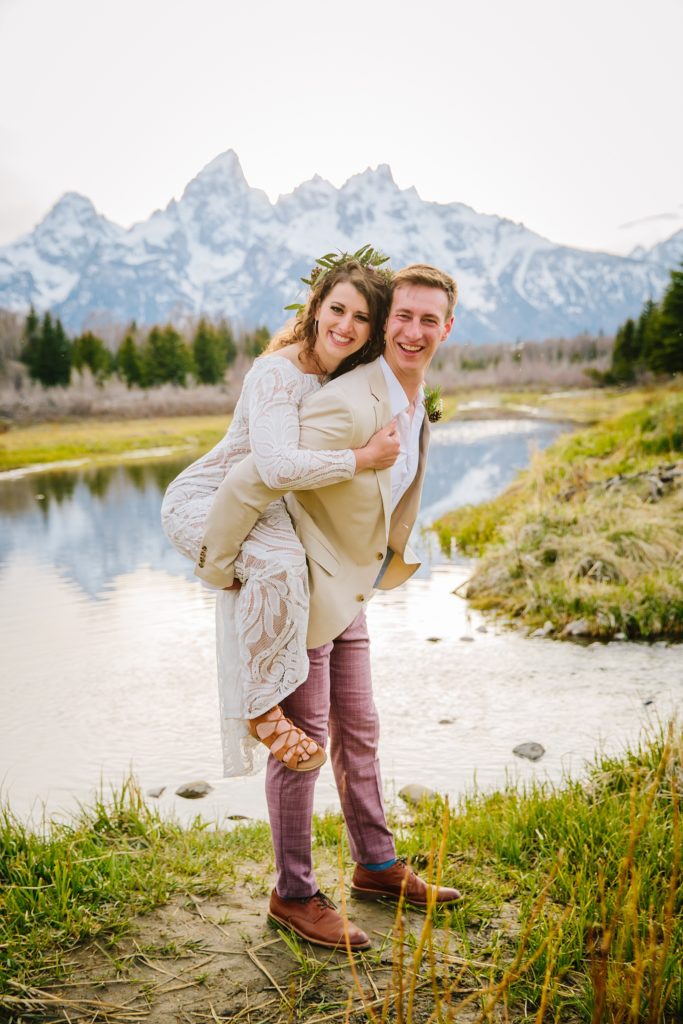 Jackson Hole wedding photographer captures groom giving bride a piggy back ride in the moutnains as the bride wears her lace wedding dress