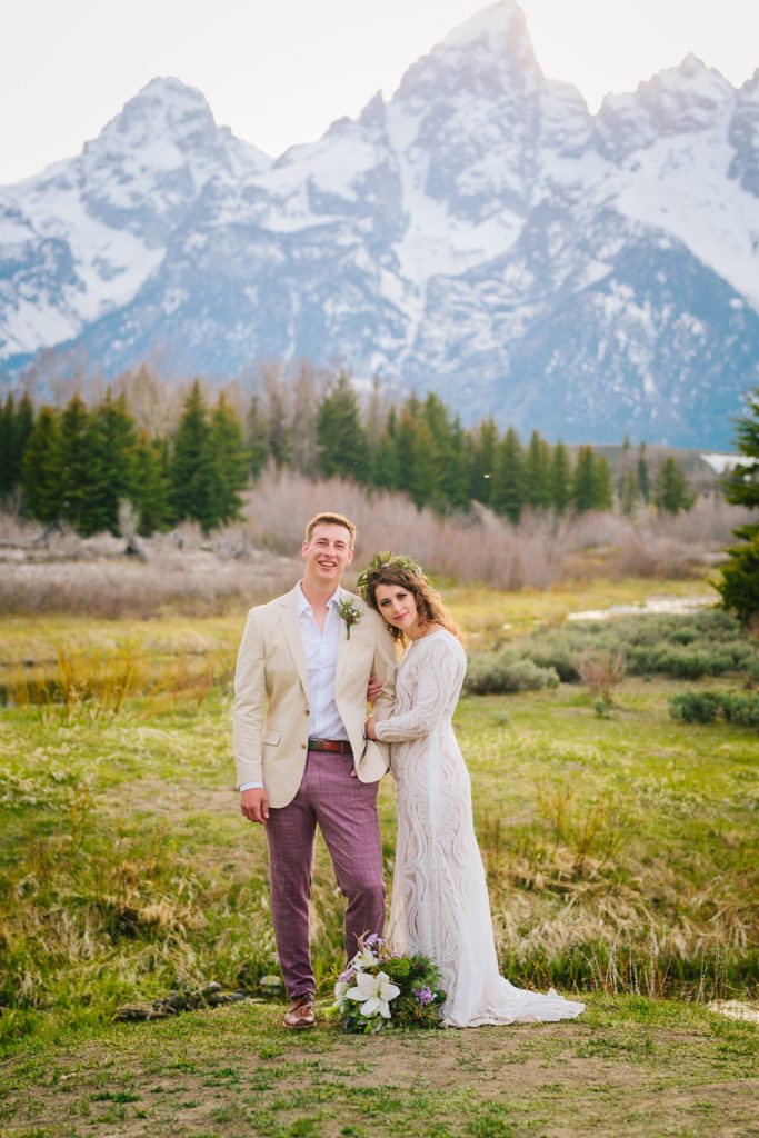 bride and groom stand in front of the grand tetons looking at the camera the brides flowers on the ground. 