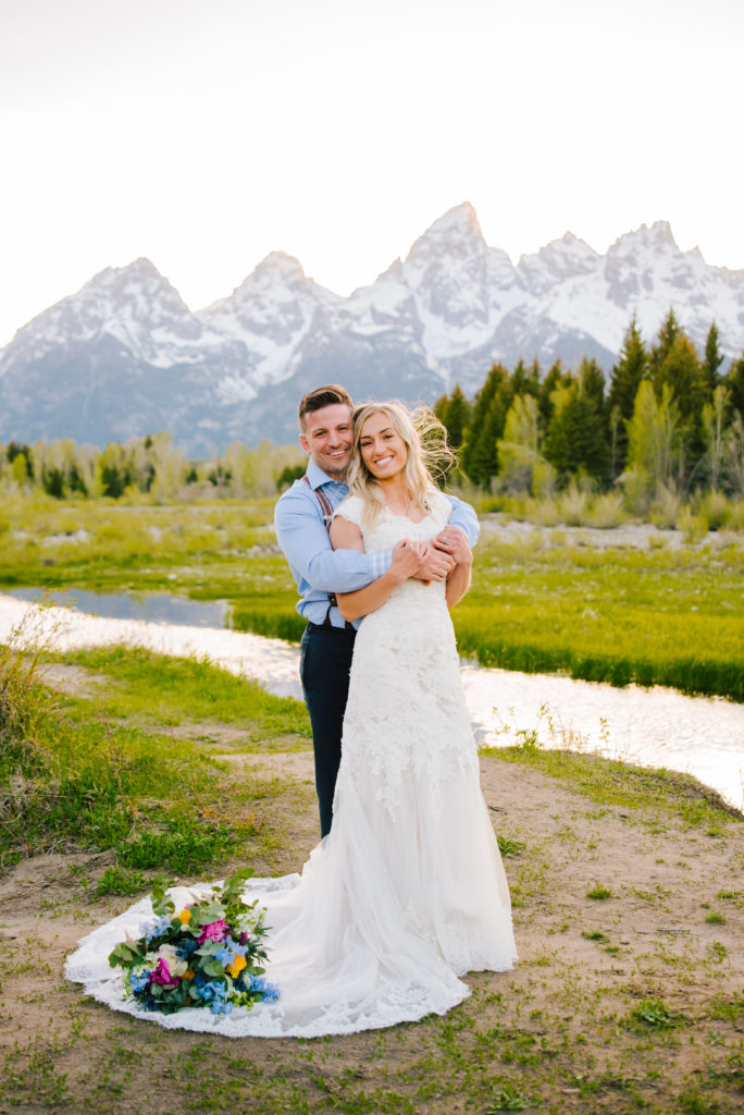 Jackson Hole wedding photographer captures grand Tetons in the distance while bride and groom embrace each other with the brides florals on the ground next to her wedding dress captured by Jackson Hole wedding photographer