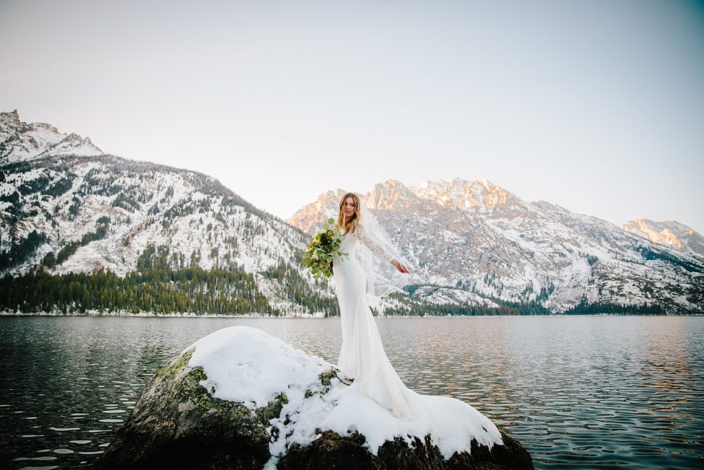young bride in a lace wedding dress stands on a rock while holding her bridal bouquet with a lake and mountains behind her 
