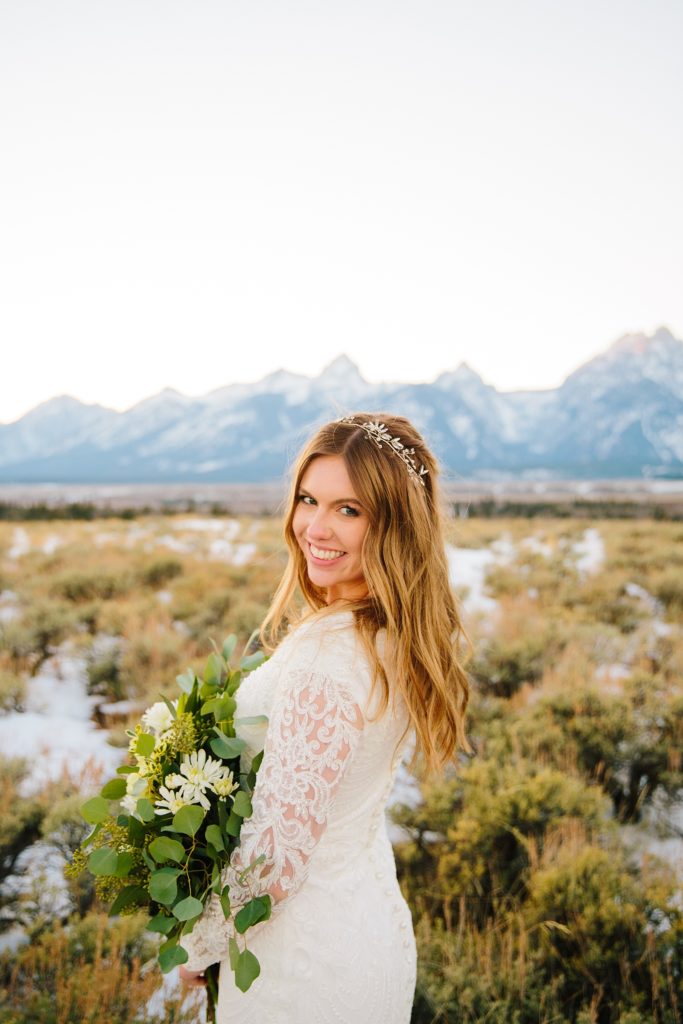 Jackson Hole wedding photographer captures  bride in a lace wedding dress holding a white floral and greenery bouquet while smiling over her shoulder