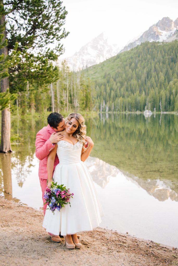 Jackson Hole wedding photographer captures groom wearing pink suit kissing bride's shoulder
