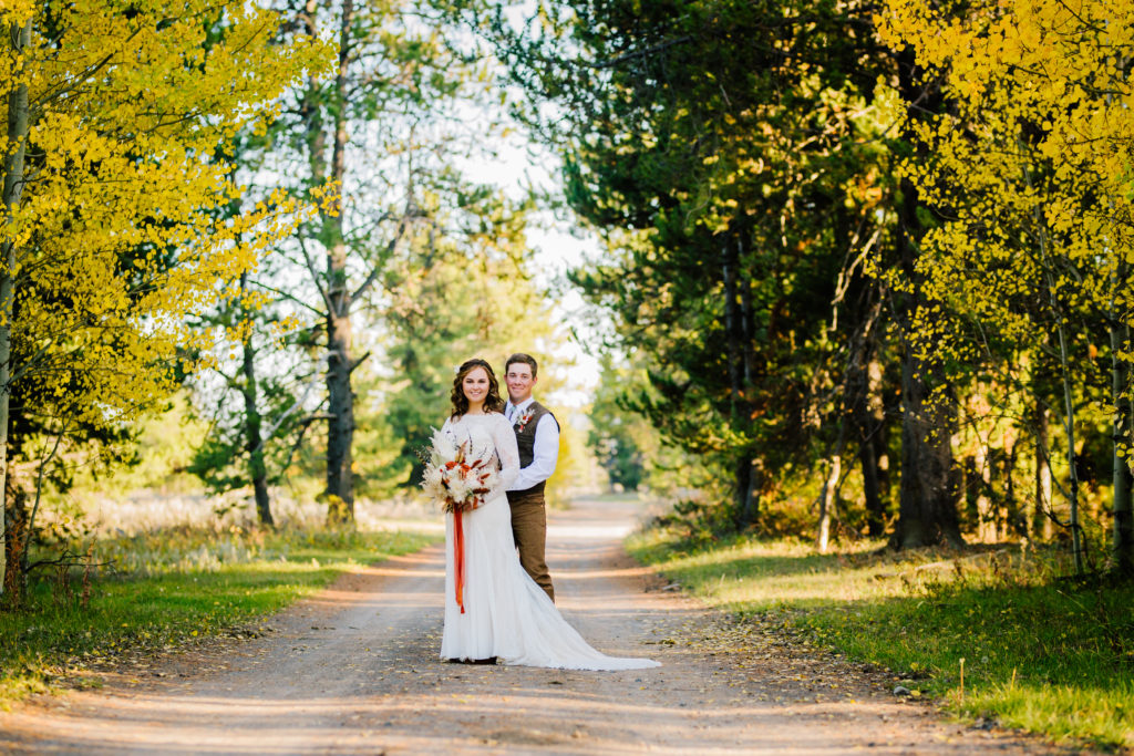 bride and groom embracing each other on a dirt path surrounded by colored trees for their fall wedding