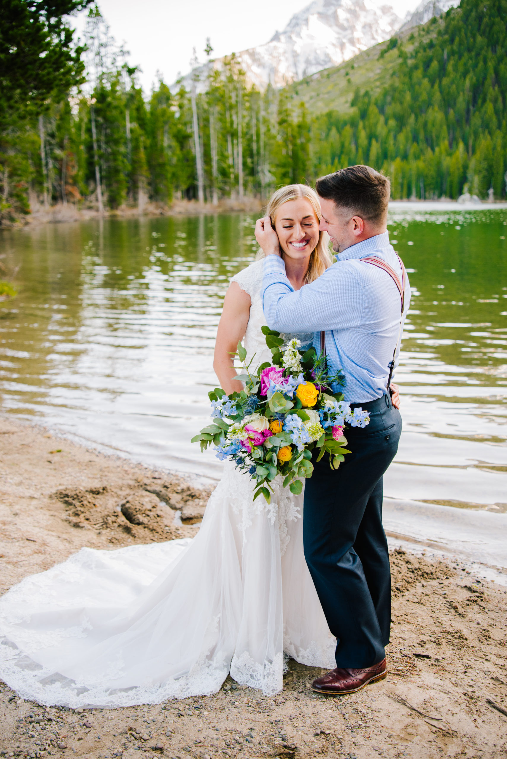 bride and groom embracing in front of a lake as the bride laughs and the groom caresses her face