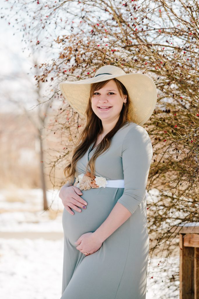 Jackson Hole wedding photographer captures woman wearing gray dress holding baby bump and wearing a hat during maternity pictures