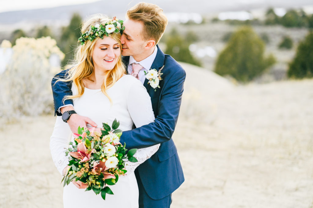 outdoor wedding captured by Pocatello wedding photographer with groom embracing bride as he stands behind her and whispers in her ear