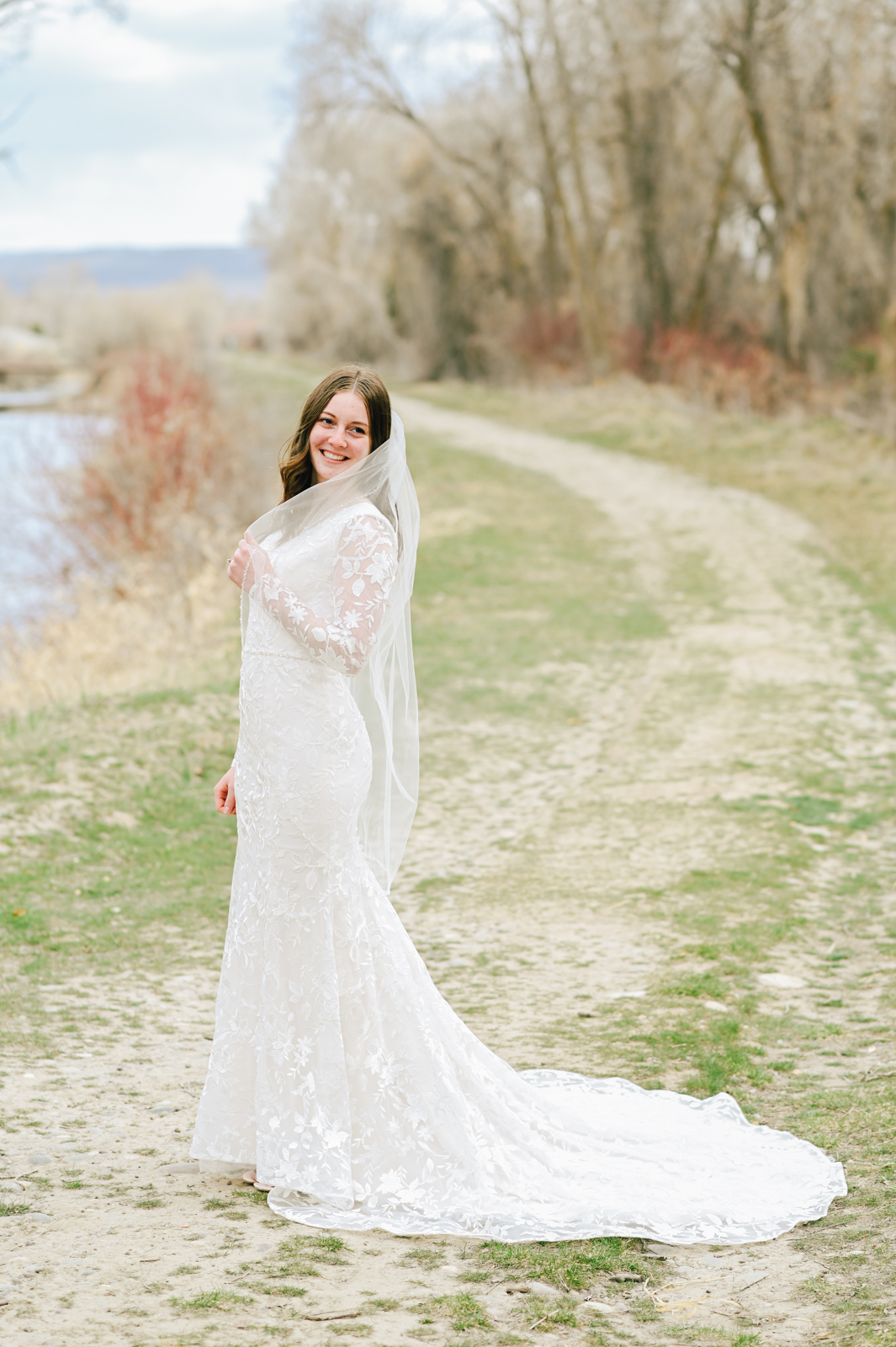 bridal portrait of bride on a dirt trail in front of a river holdher her veil as her wedding dress train falls behind her 