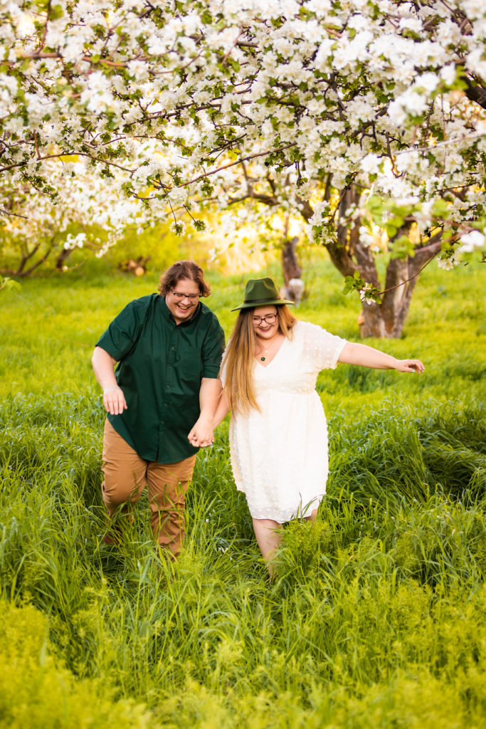 engaged couple walking through apple blossoms wearing white dress and green shirt