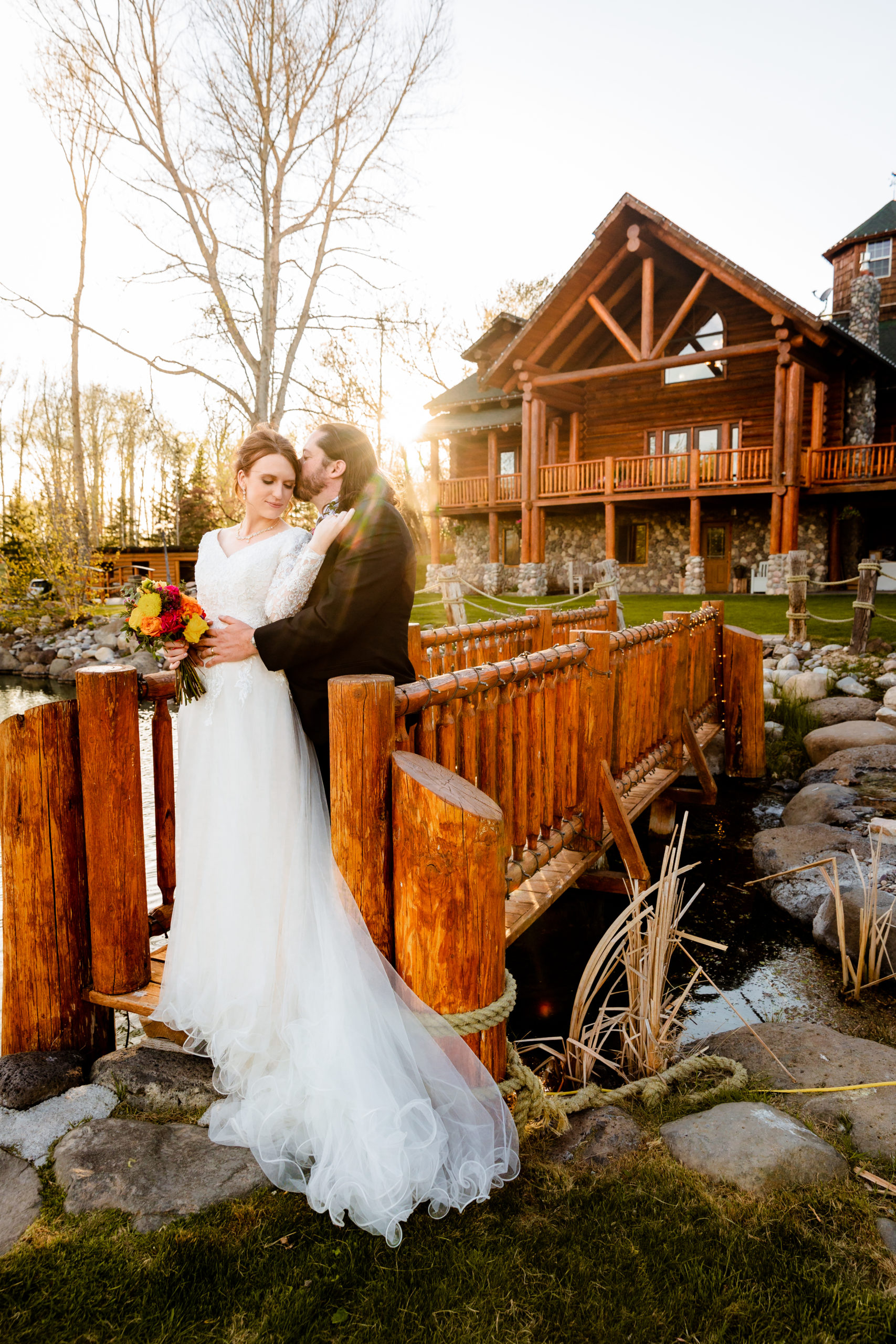 outdoor bridals at LaBelle Lake wedding venue in Rigby Idaho with groom embracing bride as he stands behind her and the sun sets by the cabin