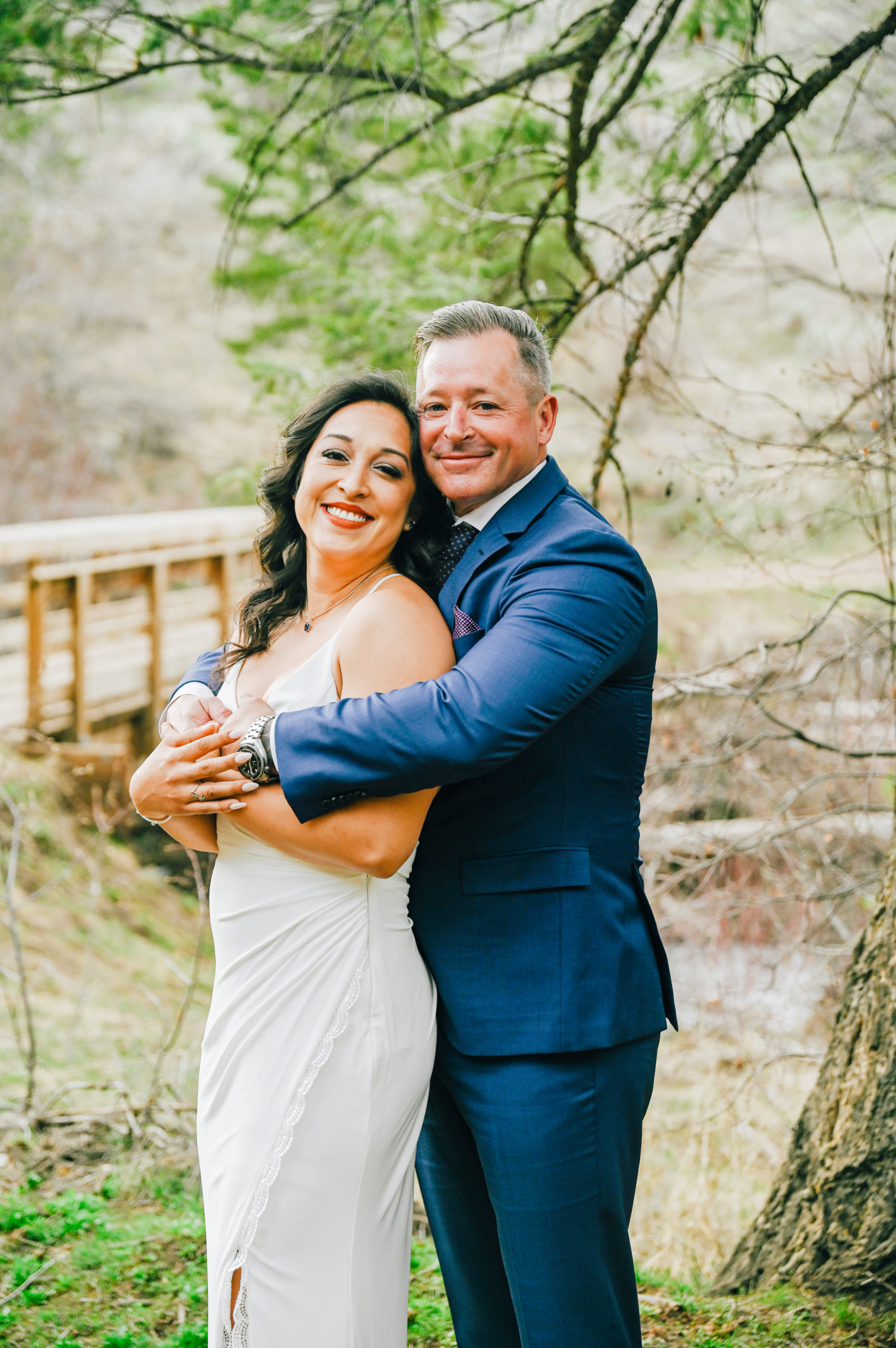 groom standing behind the bride and wrapping his arms around her shoulder with a bridge and forest behind them