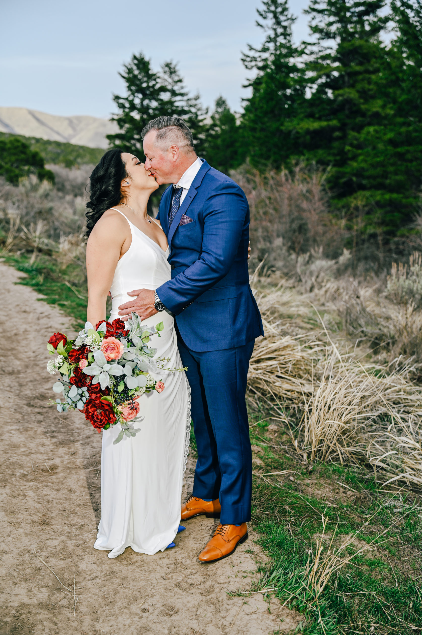 bride holding her red florals to her side as her groom holds her waist and kisses her captured by Pocatello wedding photographer