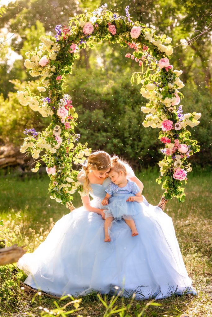 bride in blue dress sits on swing covered in pink flowers with toddler daughter embracing