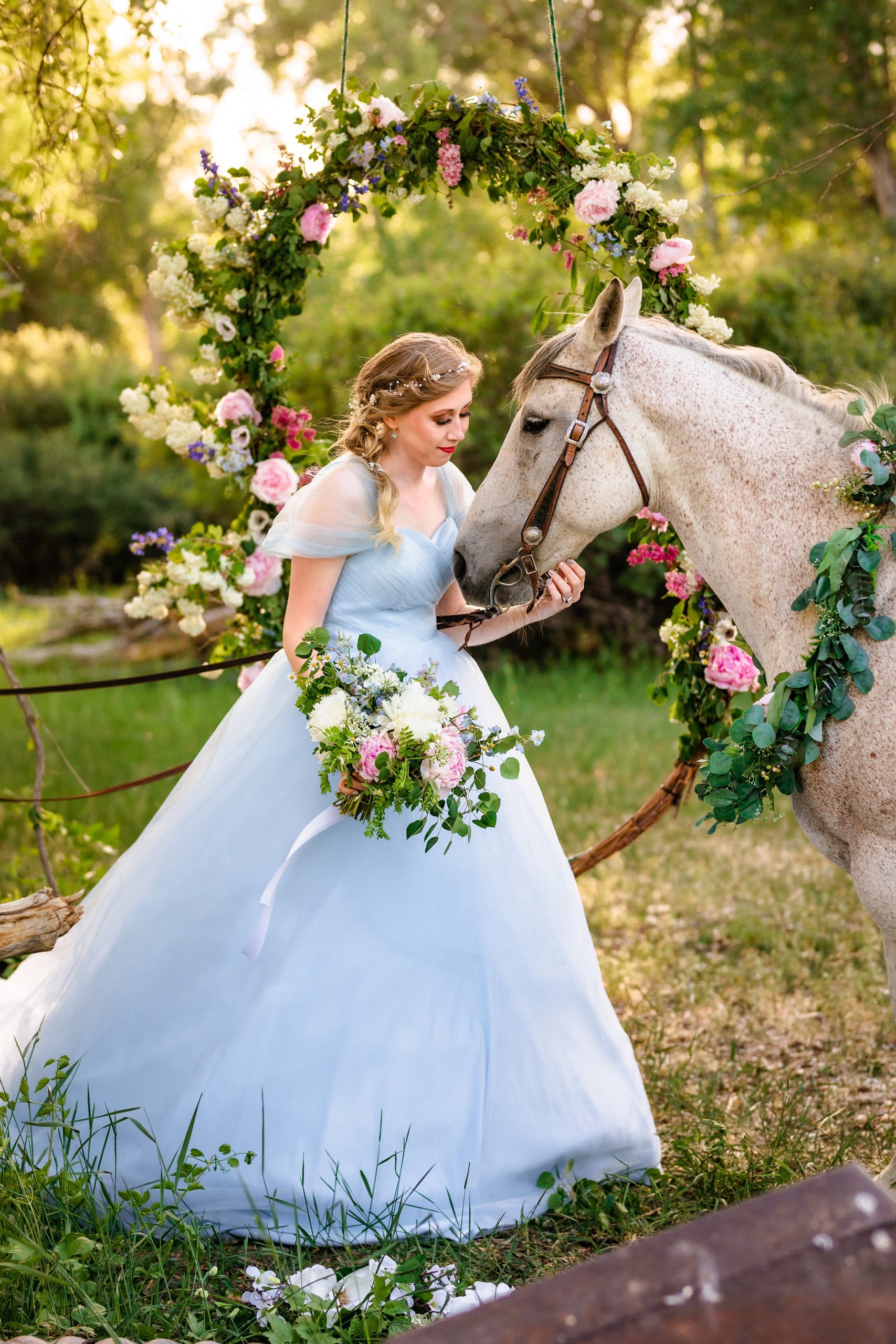 bride in blue dress reaches for white horse with flowers around neck