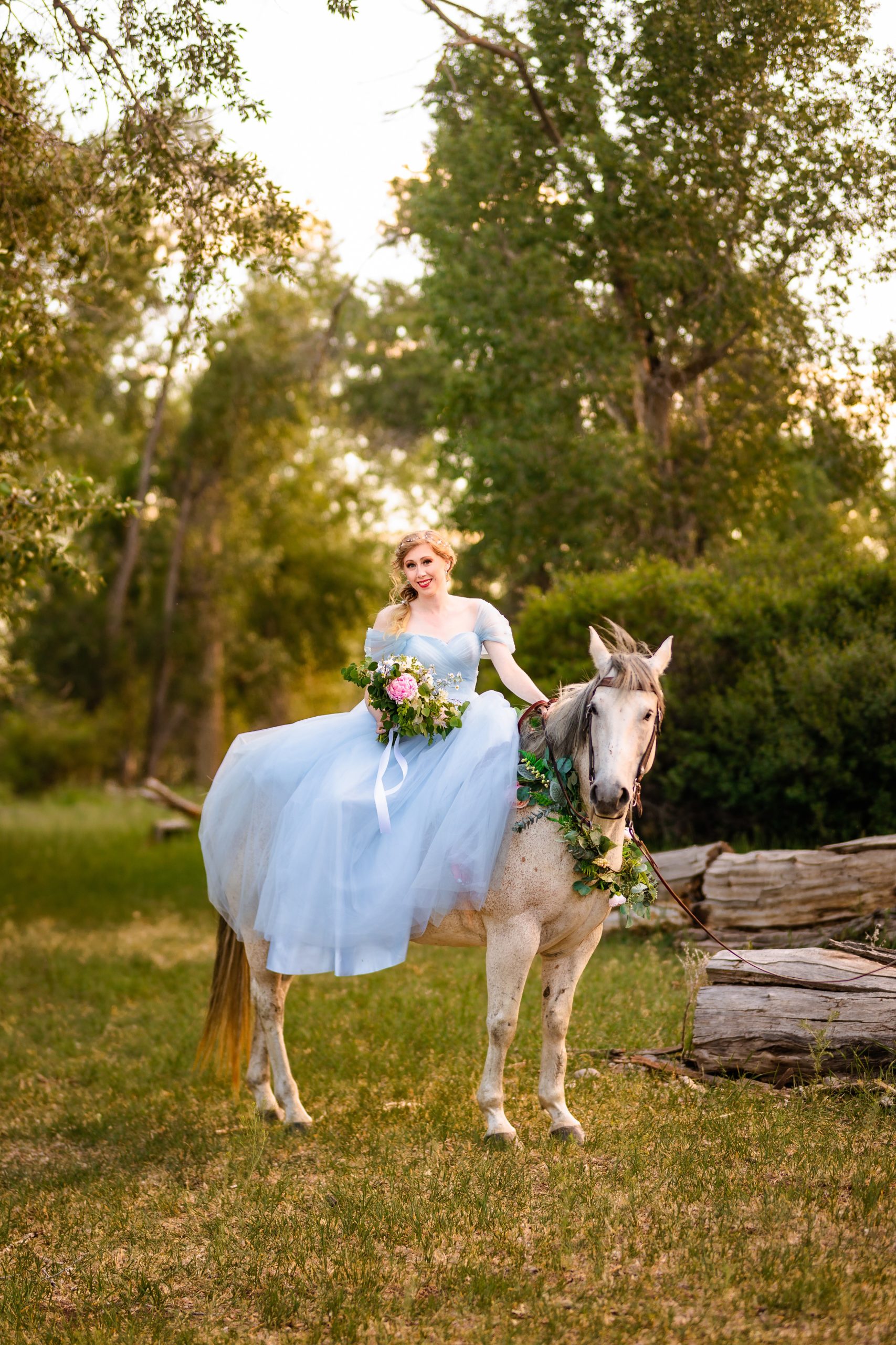princess bride in blue dress sits on white horse in idaho forst