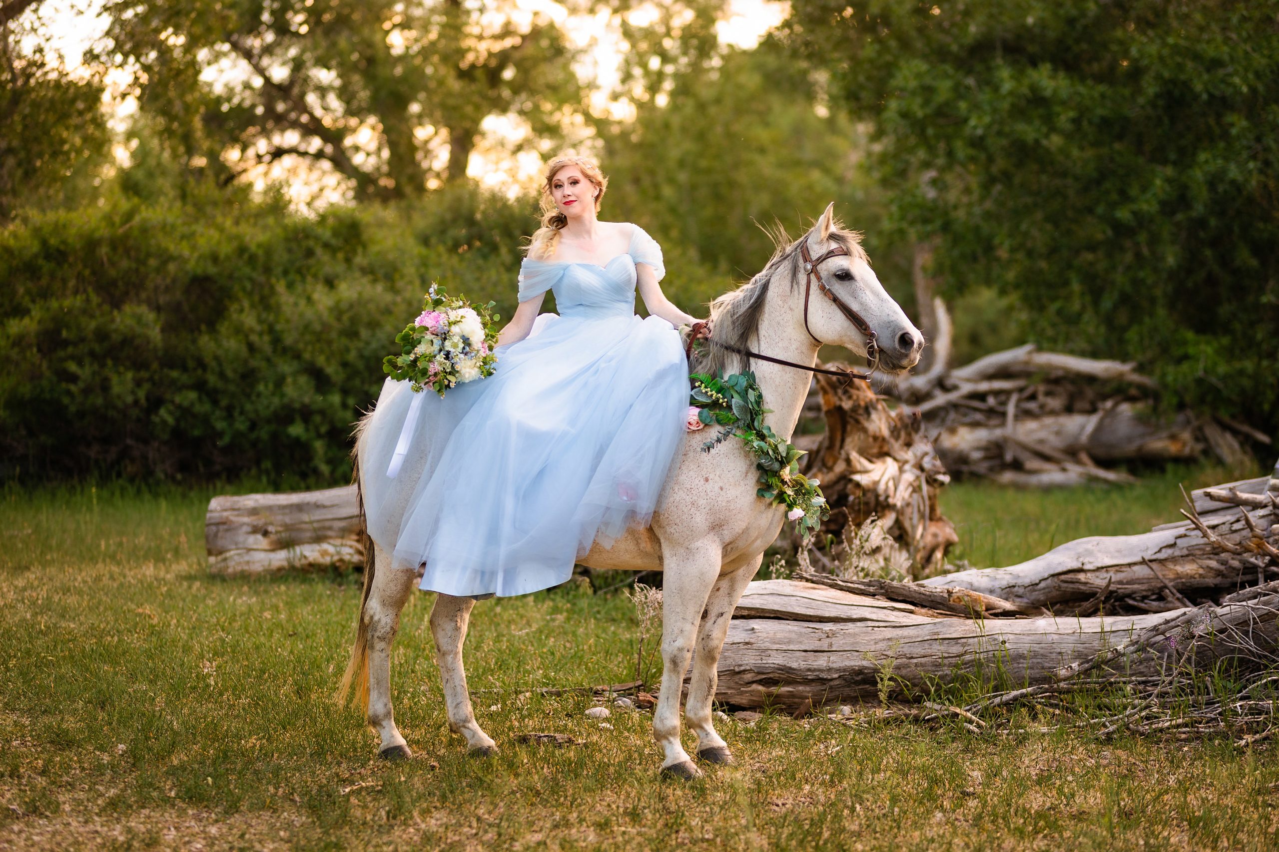 bride in blue wedding dress sitting on horse with flowers draped in one hand while looking at the camera