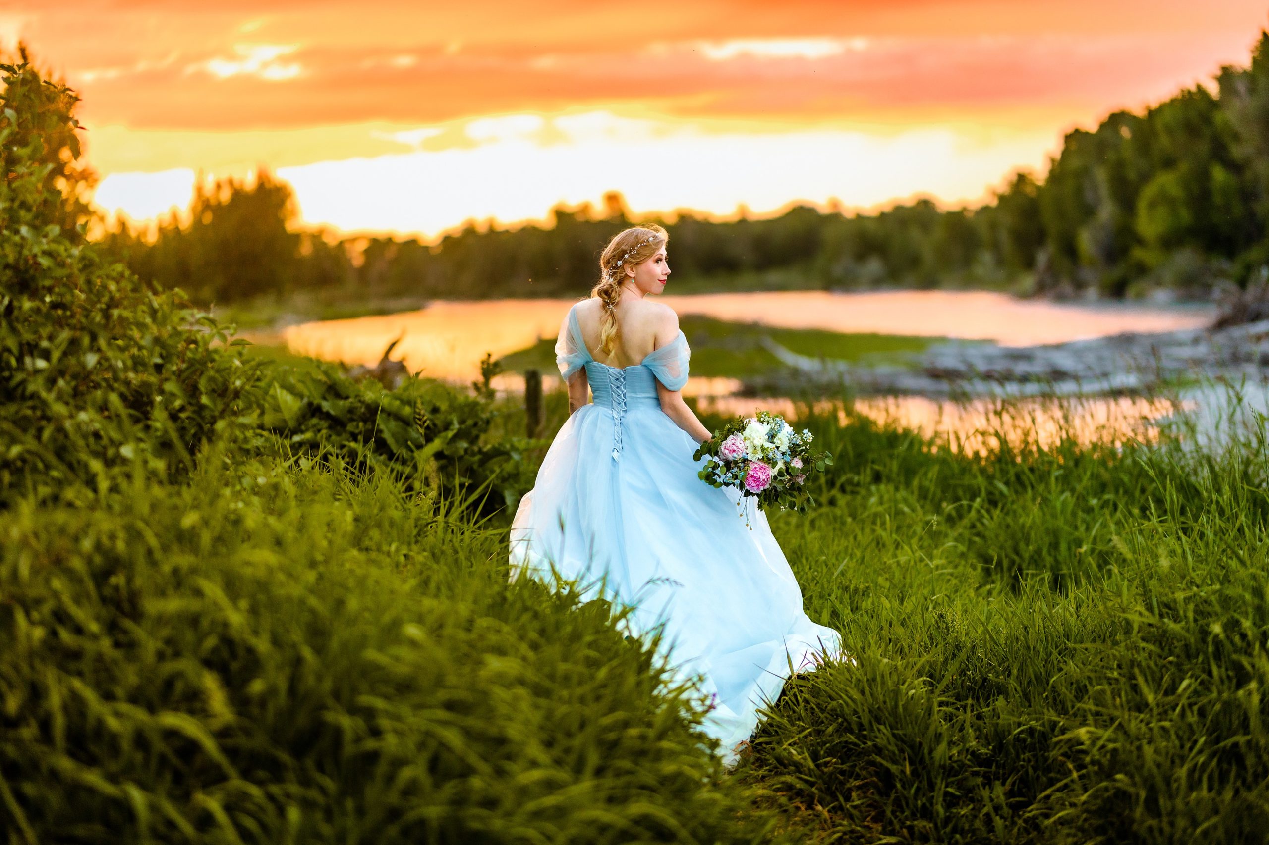 golden bride in blue dress with orange and pink sunset next to beautiful river