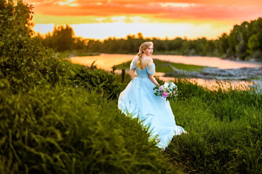 bride in blue dress with orange and pink sunset next to beautiful river