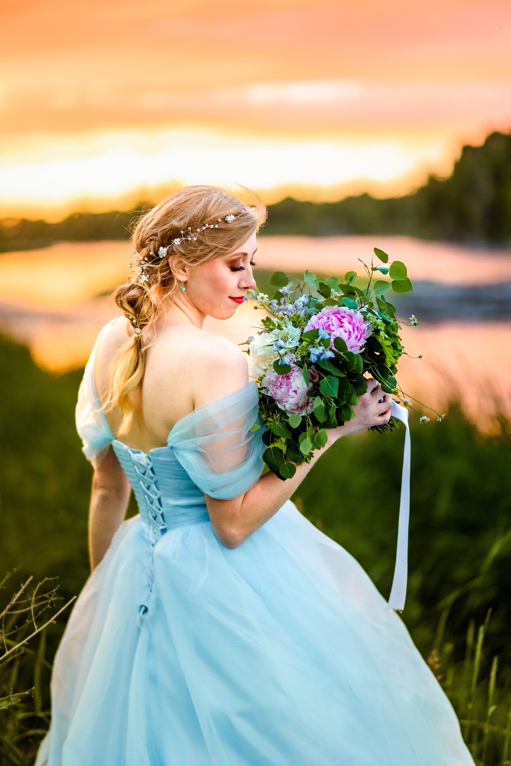 smelling flowers bride in blue dress with orange and pink sunset next to beautiful river