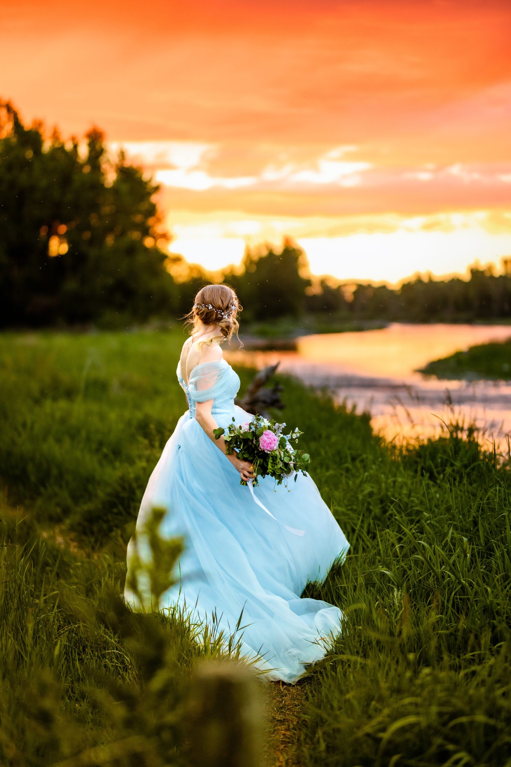 back to camera bride in blue dress with orange and pink sunset next to beautiful river
