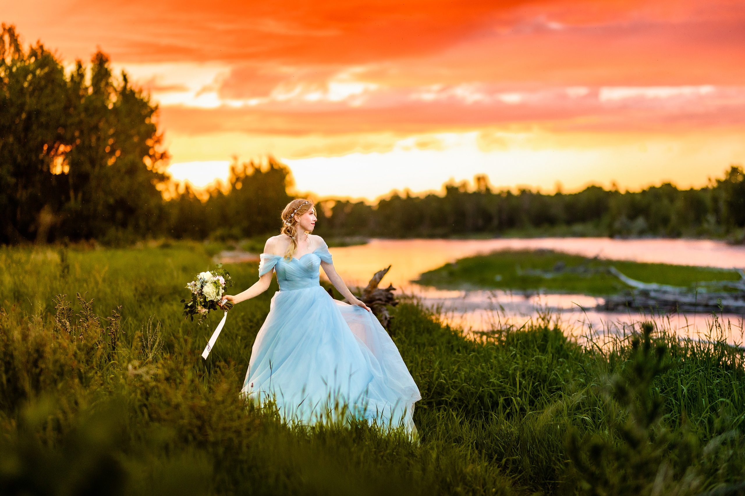 windy bride in blue dress with orange and pink sunset next to beautiful river
