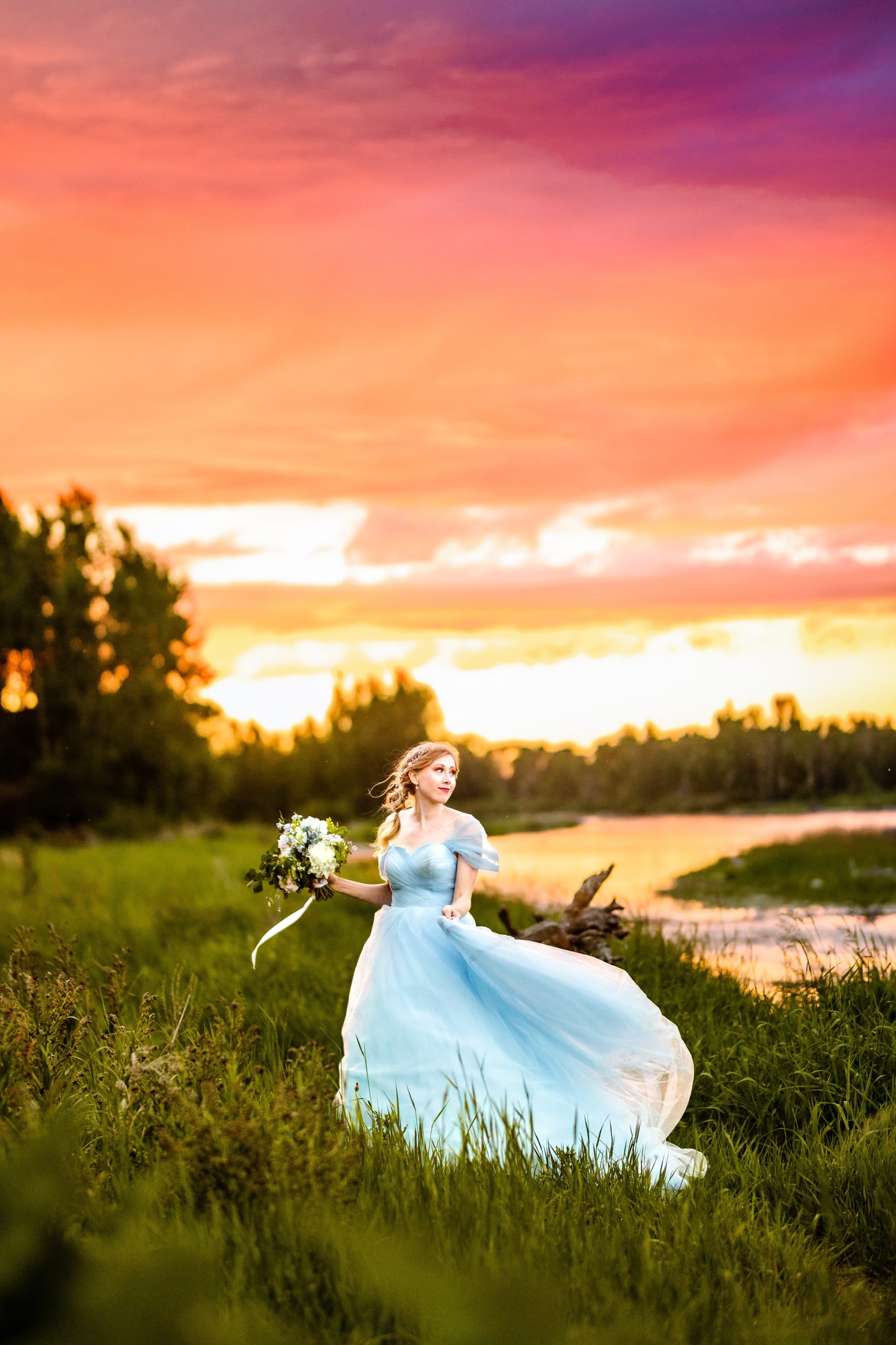 bride in blue dress with orange and pink sunset next to beautiful river swishing her dress