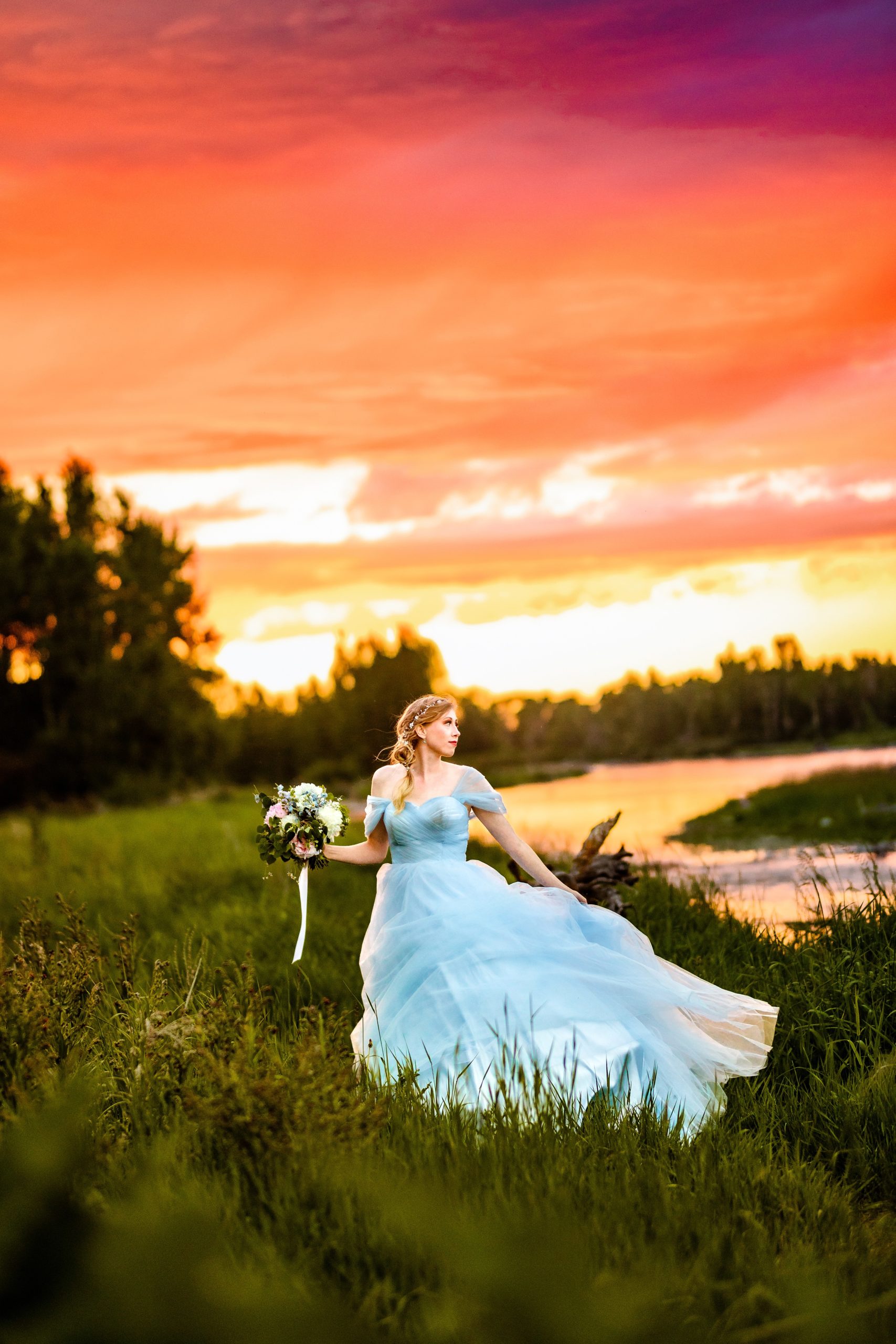 wind blows bride in blue dress with orange and pink sunset next to beautiful river