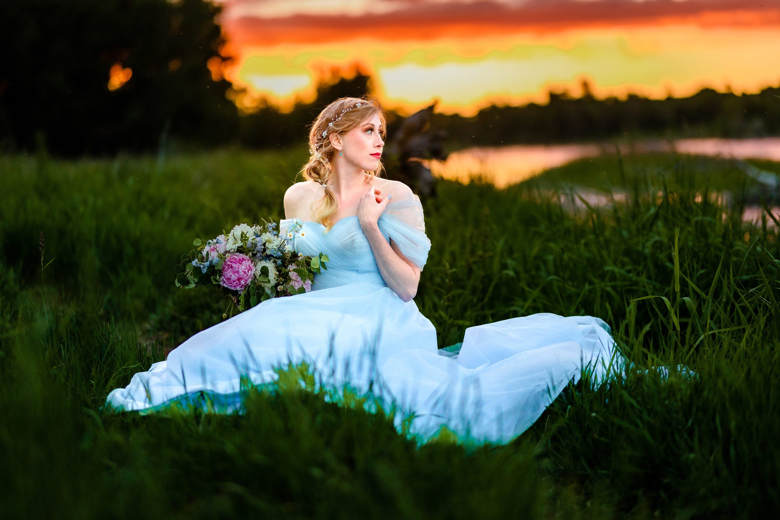 sitting bride in blue dress with orange and pink sunset next to beautiful river