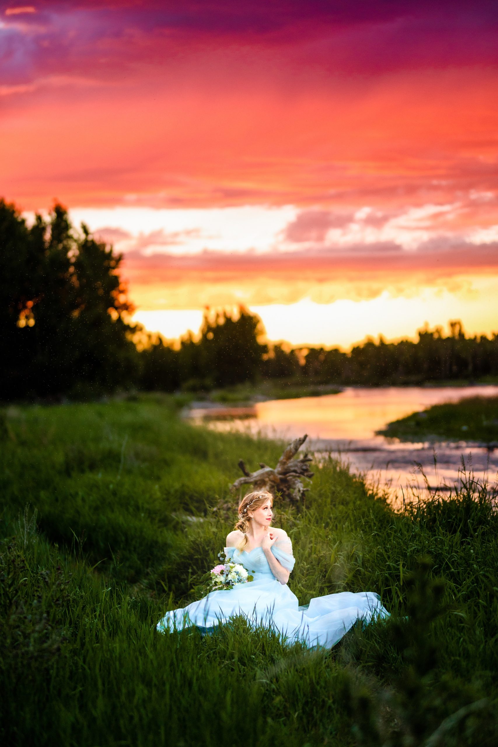 bride in blue dress with orange and pink sunset next to beautiful river sitting and gazing