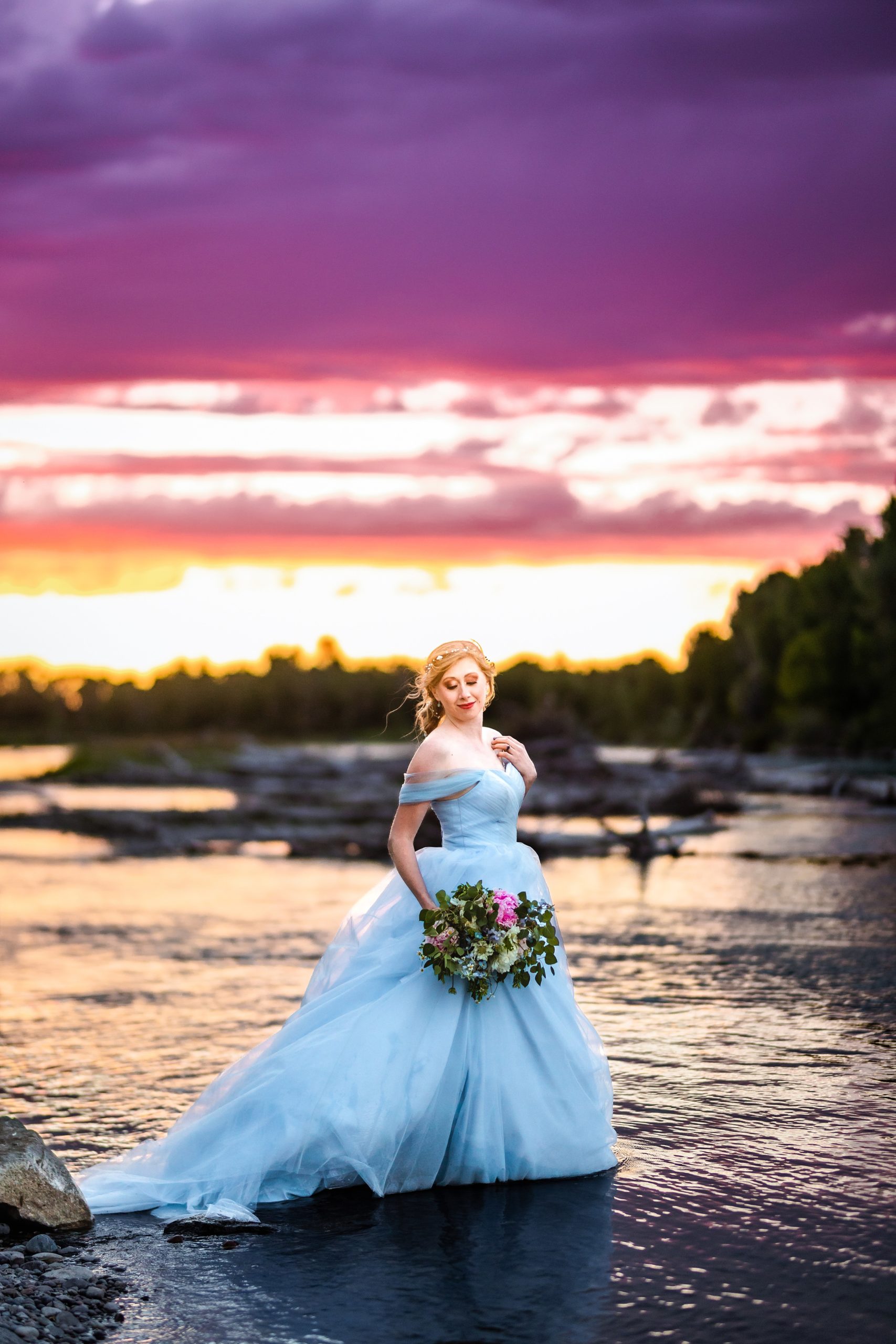 standing in river bride in blue dress with orange and pink sunset next to beautiful river