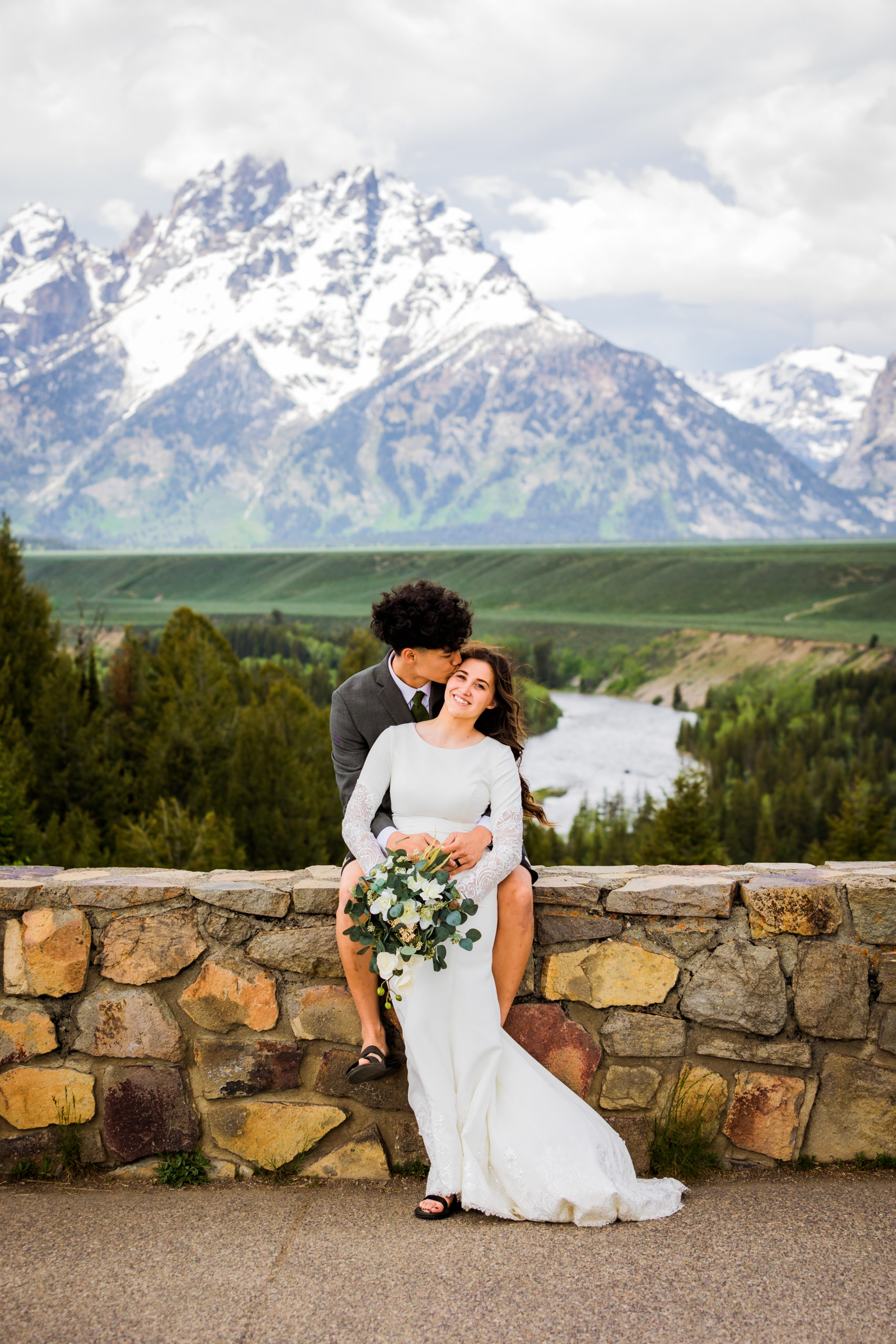 groom sitting on a stone wall while holding his bride from behind as she smiles and the mountains stand behind them 
