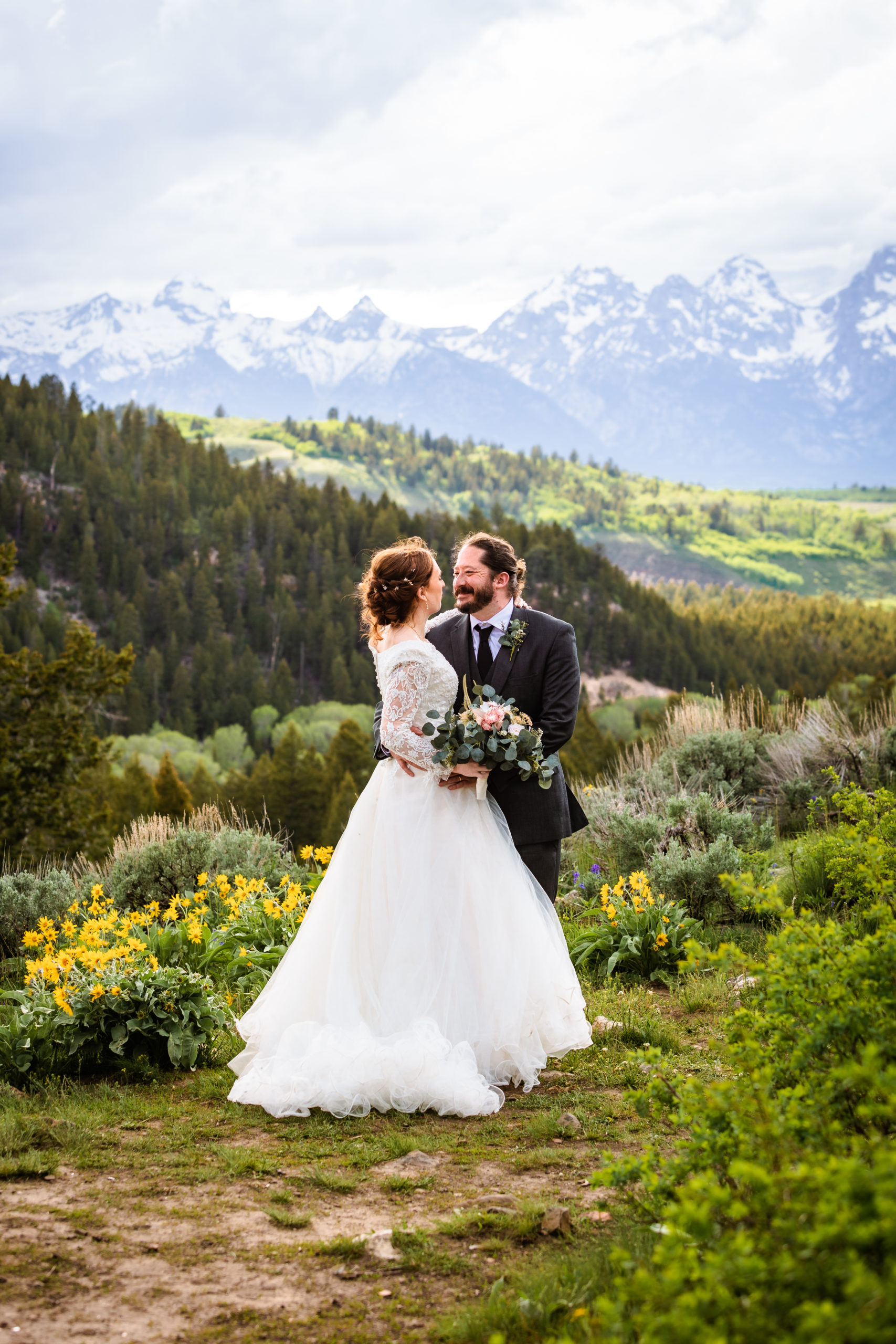 bride and groom gaze into eachother eyes while standing in front of the tetons in grant teton national park surrounded by lush green and yellow wild flowers