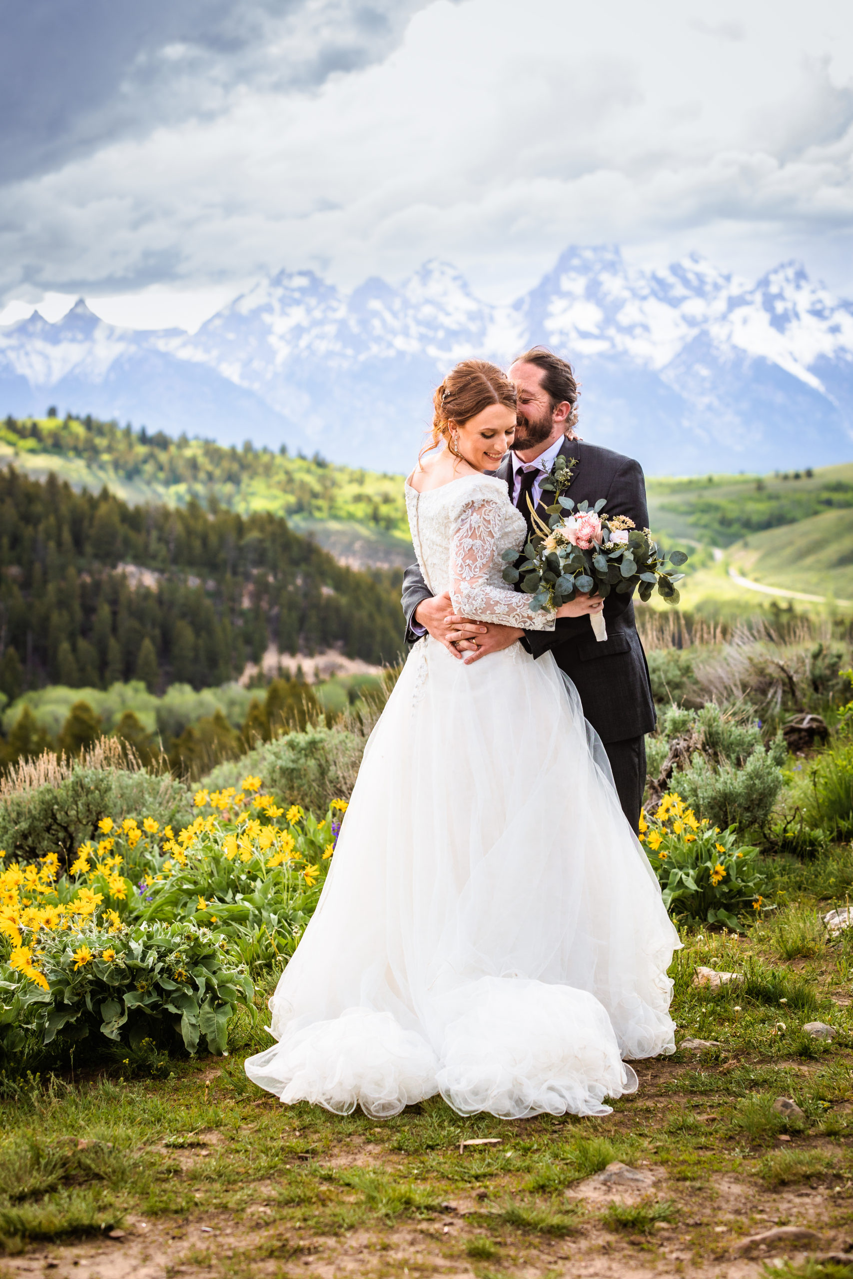 bride looks at her shoulder while groom lightly kisses her on the head they are surrounded by wildflowers that are yellow