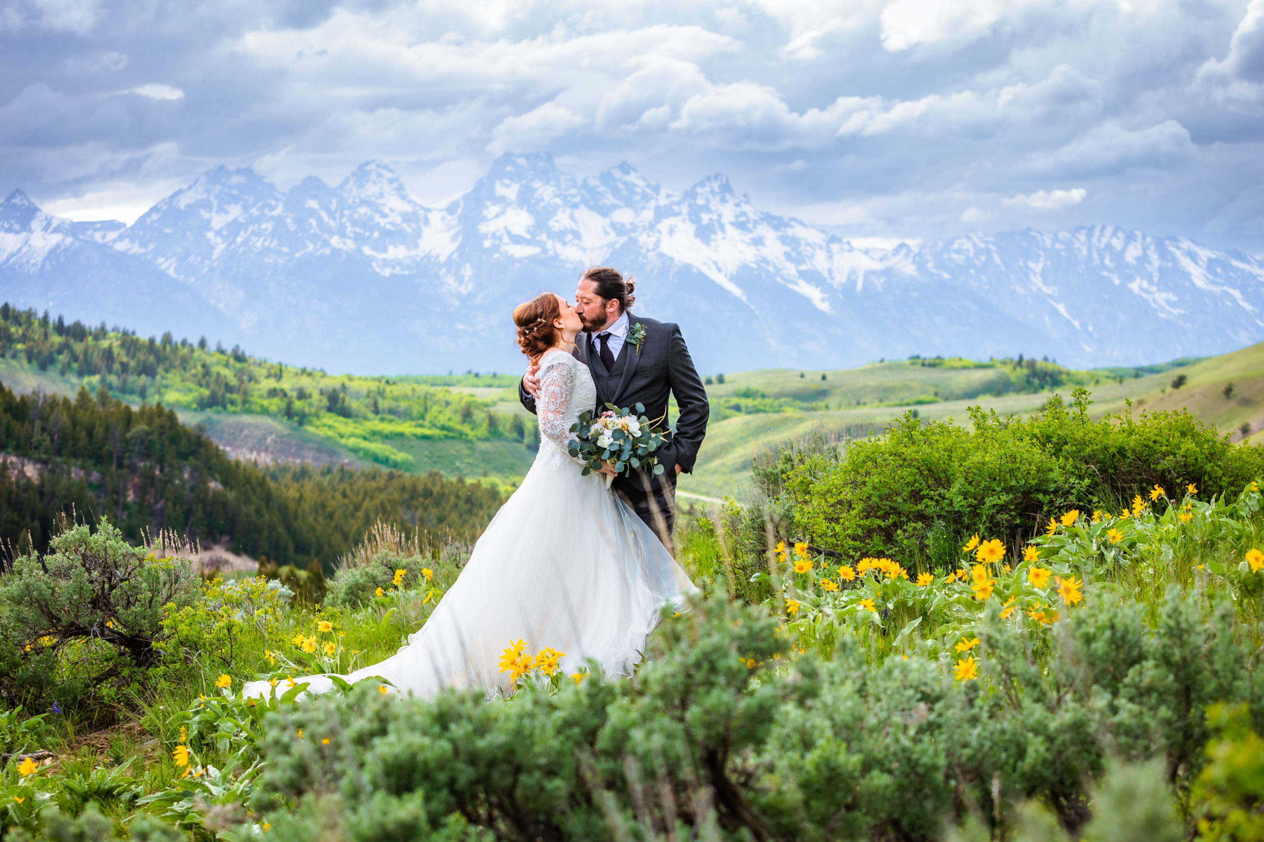 bride and groom kiss in a valley with the tetons behind them the grass is green with wild flowers