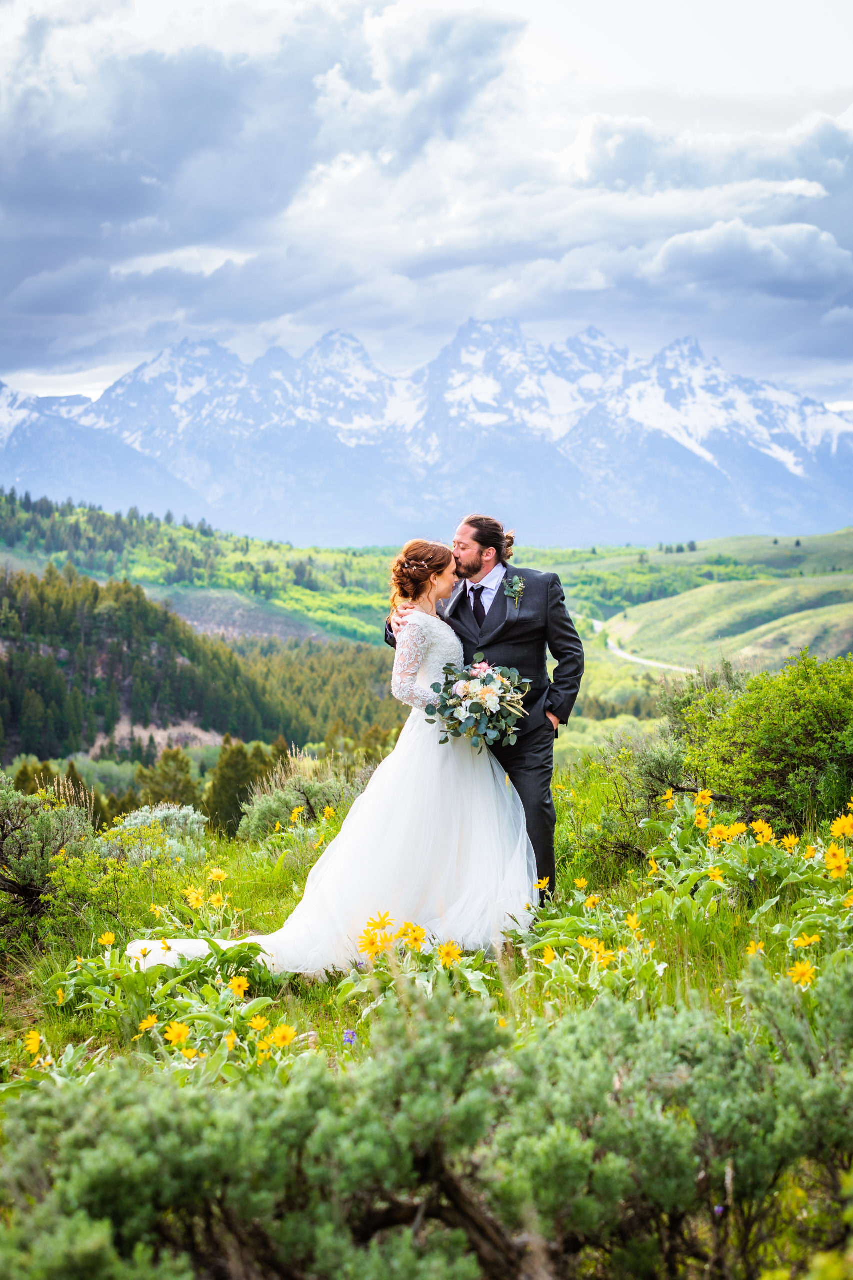 groom kisses bride on head while standing with tetons behind them the grass is green and full of wild flowers