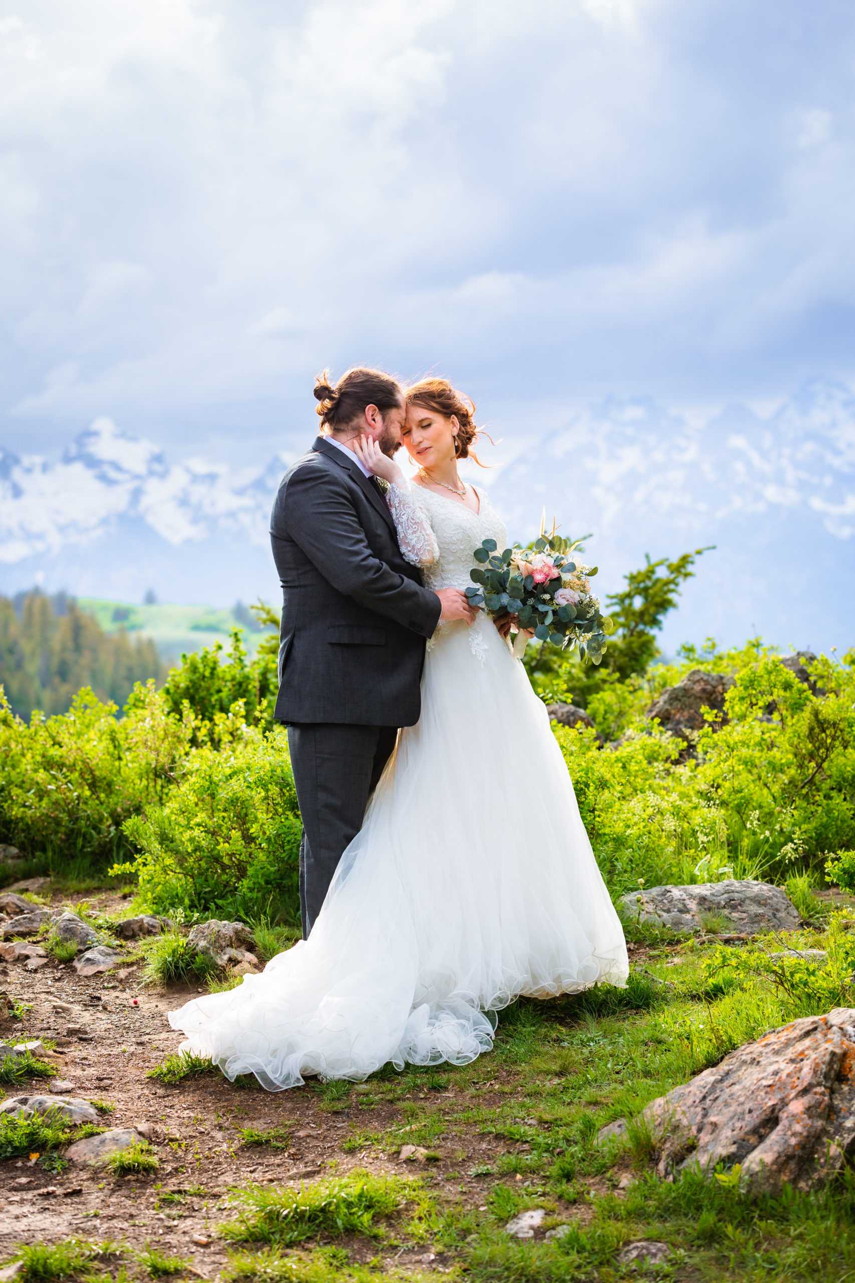 bride reaches for groom to be closer to her as they stand in green grass with the tetons behind them