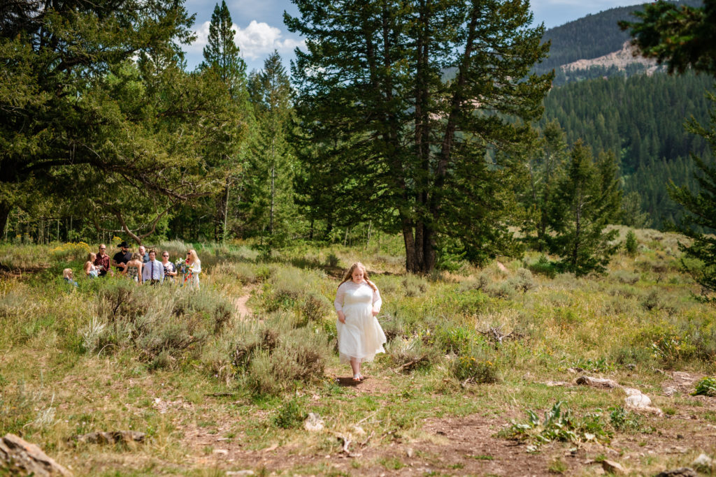 Jackson Hole wedding photographers capture bride walking through grassy area to Windy Wedding Tree Elopement