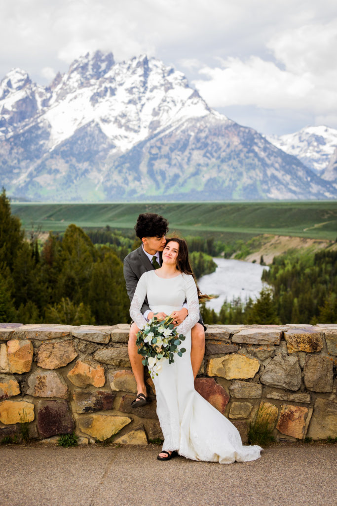 Jackson Hole wedding photographer captures bride and groom sitting on stone wall during portraits