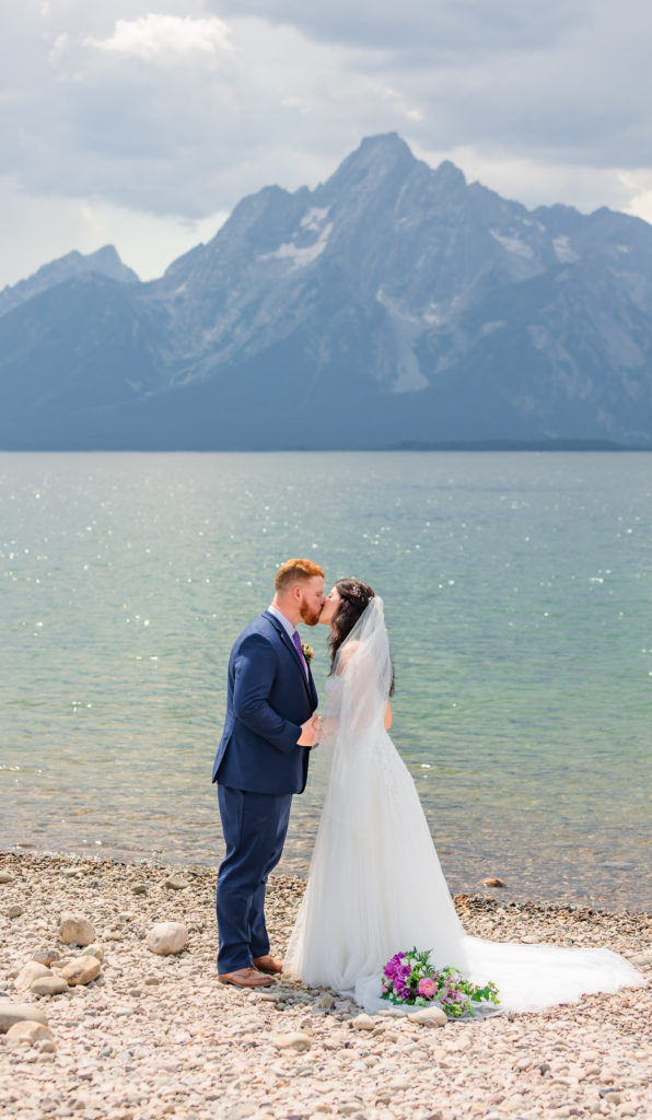Jackson hole wedding photographer captures bride and groom kissing along side water in grand Teton national park