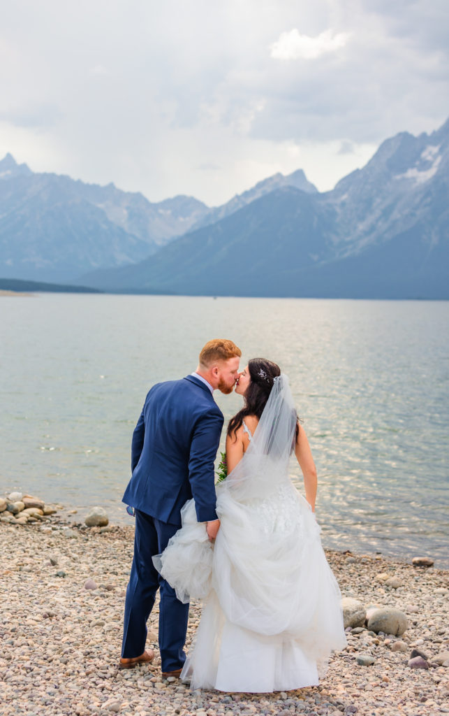 Jackson Hole wedding photographer captures bride and groom facing Colter Bay kissing while groom holds bride's train