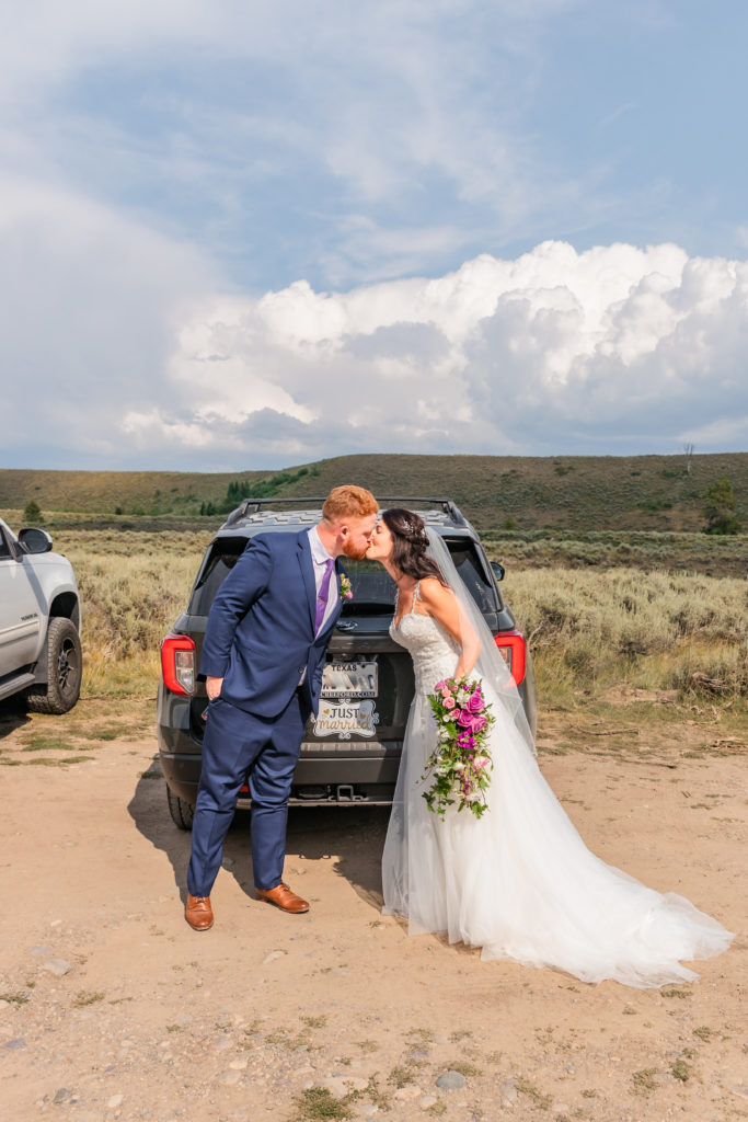Jackson Hole wedding photographers capture newly married couple wearing wedding attire kissing in front of parked car in Grand Teton National Park wedding