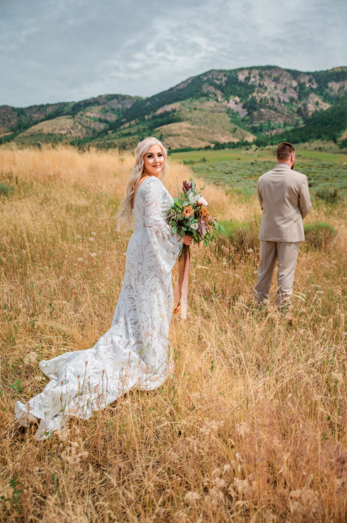 bride behind anxious groom waiting to reveal her dress while standing in the field with mountains behind them.