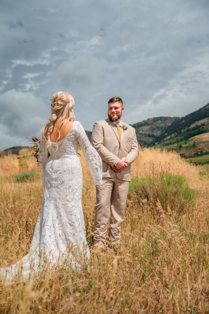 Bride reveals dress to groom. Groom smiles as the stand in a wheat colored field.
