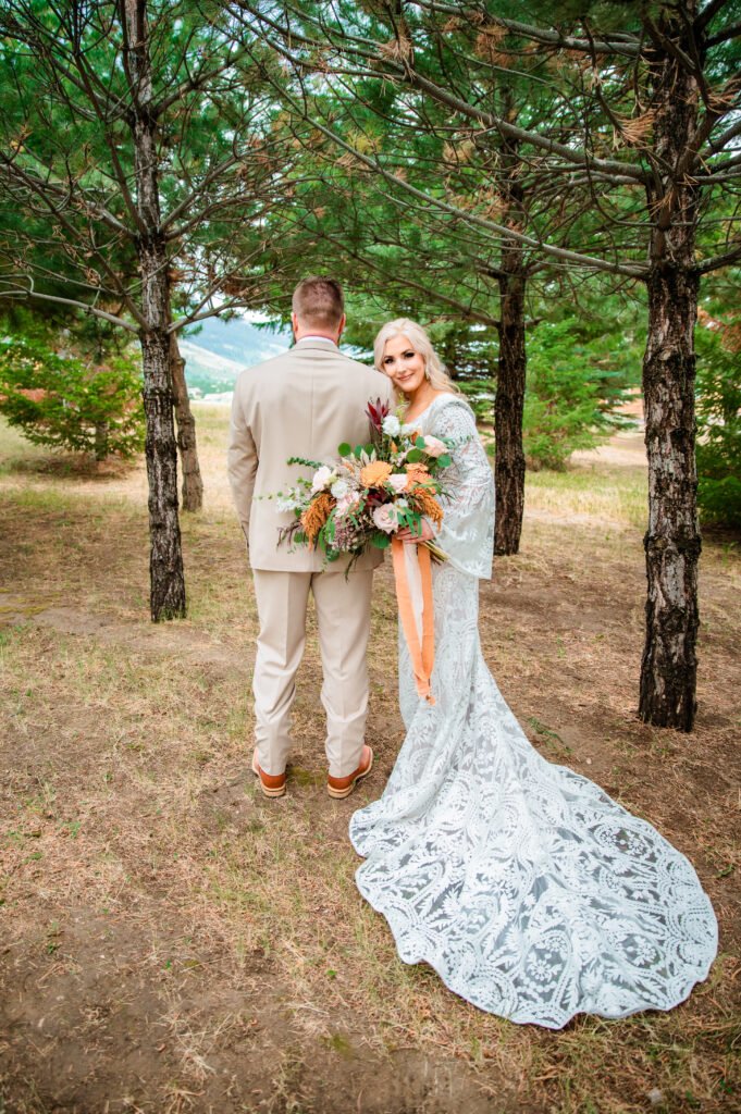 Bride shows off her lace dress and large orange and cream bouquet while groom faces away from the camera while standing in the forest.