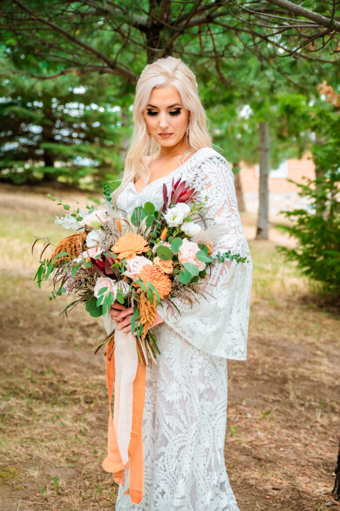 Jackson Hole bride shows off her lacey boho dress and bouquet full of orange and cream tones. Bride is looking down at her flowers